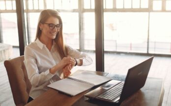 smiling young female entrepreneur checking time on wristwatch while working on laptop in modern workspace