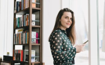 smiling young lady messaging on smartphone near window in library
